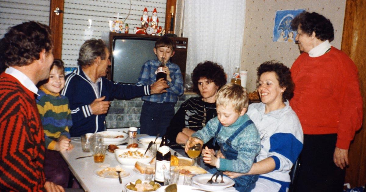 An einem gedeckten Tisch sitzt eine Familie. Ein Junge trinkt eine Cola.